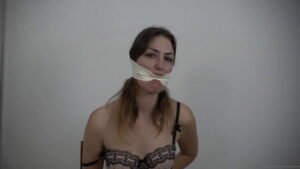 Lola on a chair with her hands tied behind her back gets gagged with microfoam tape